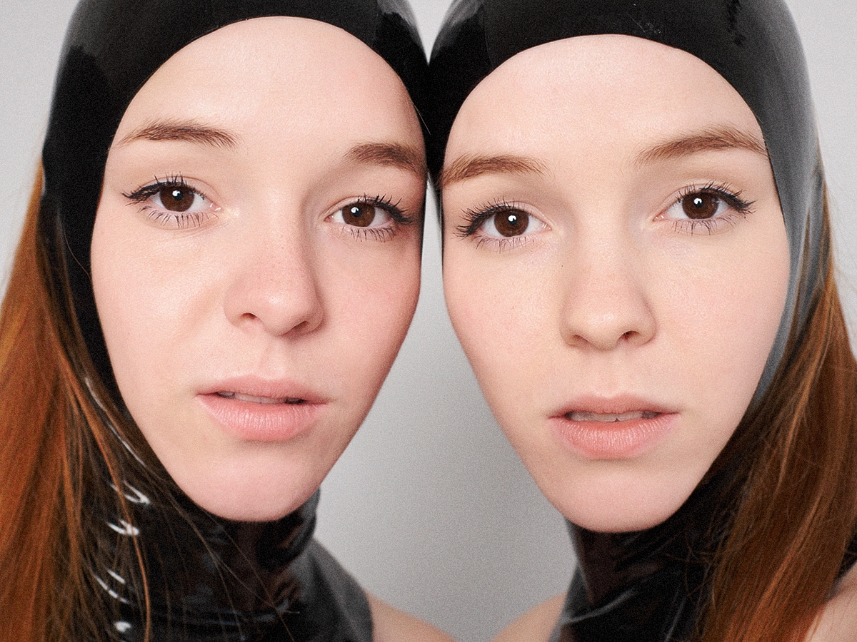 Close up photo of the Reyes Twins, Naomi & Ruth wearing black latex hoods.